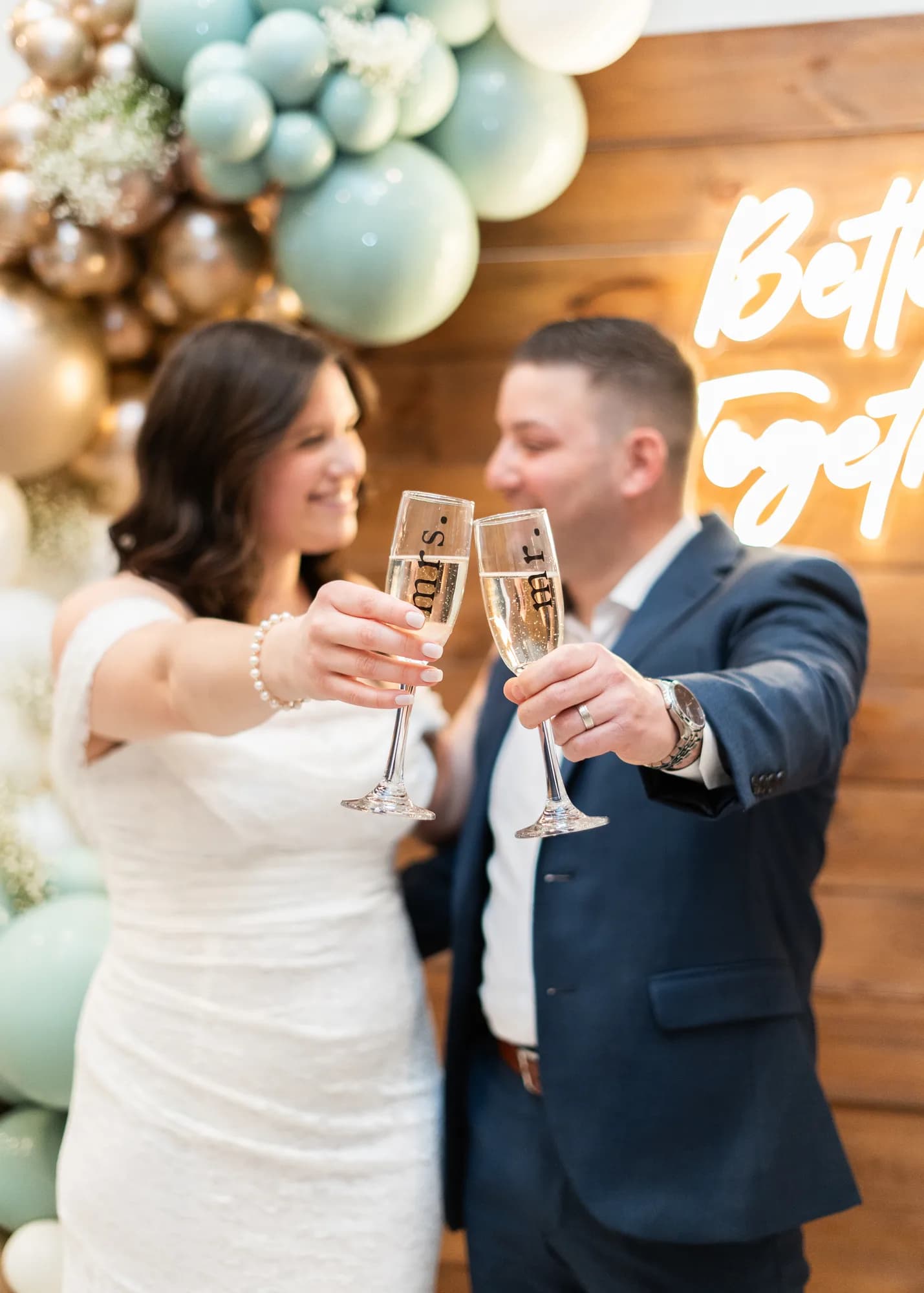 Couple toasting with personalized glasses in front of a balloon arch and neon sign