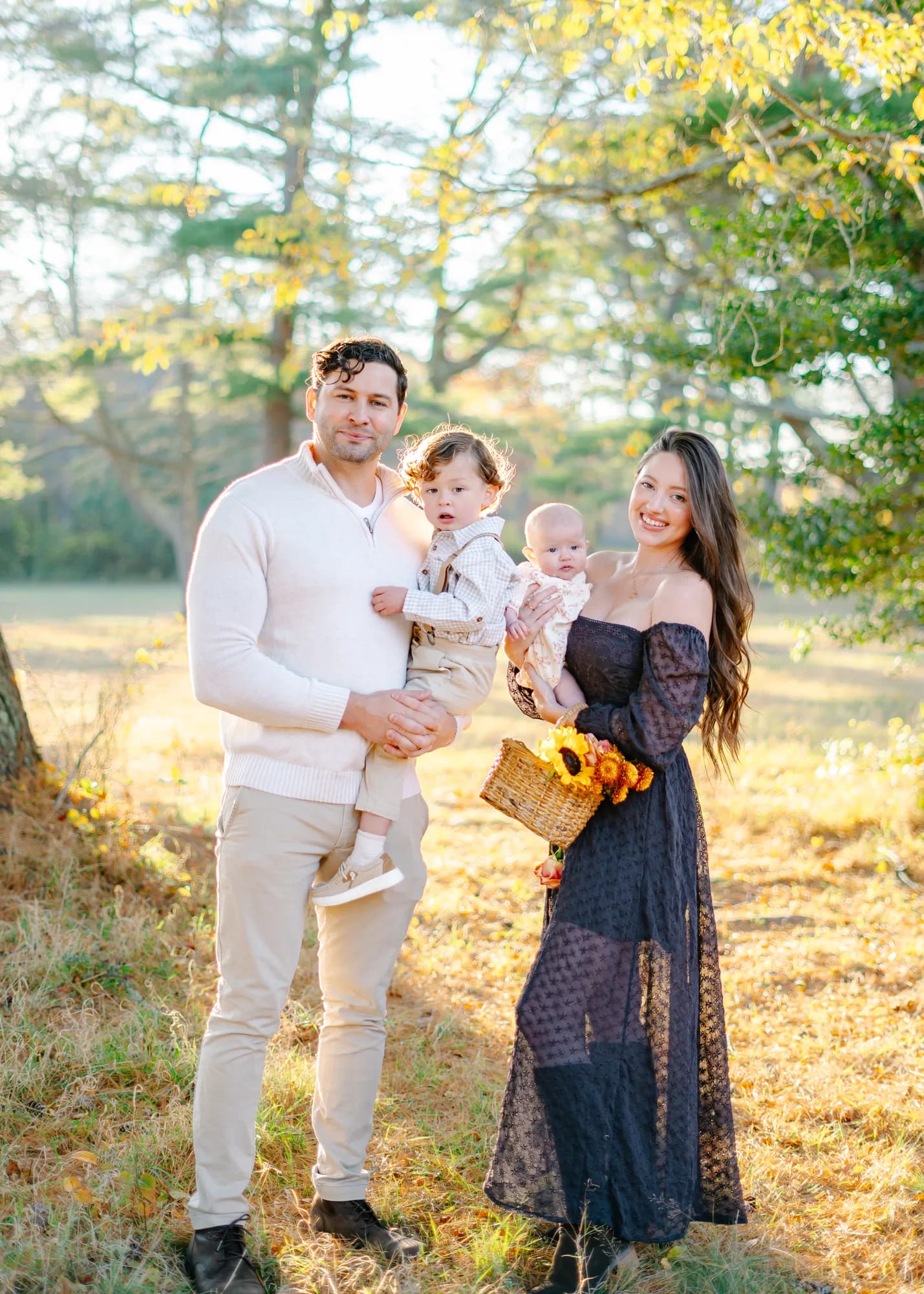 Parents holding their two young children in a golden field, a basket of sunflowers in hand