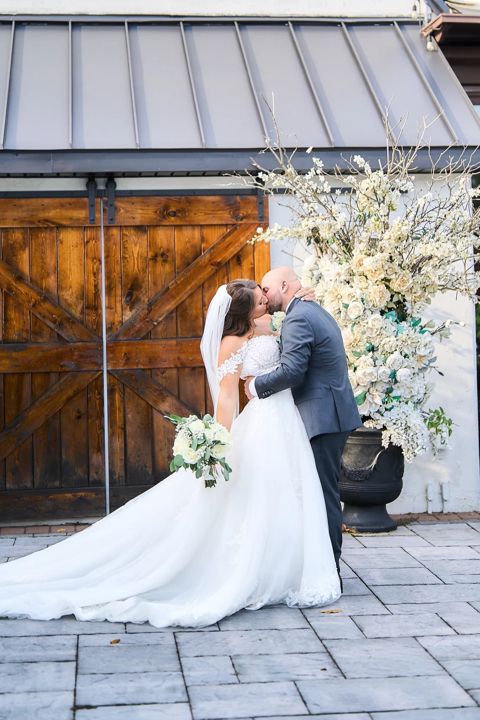 Amanda and Gabriele kissing beside the barn door and a white floral urn