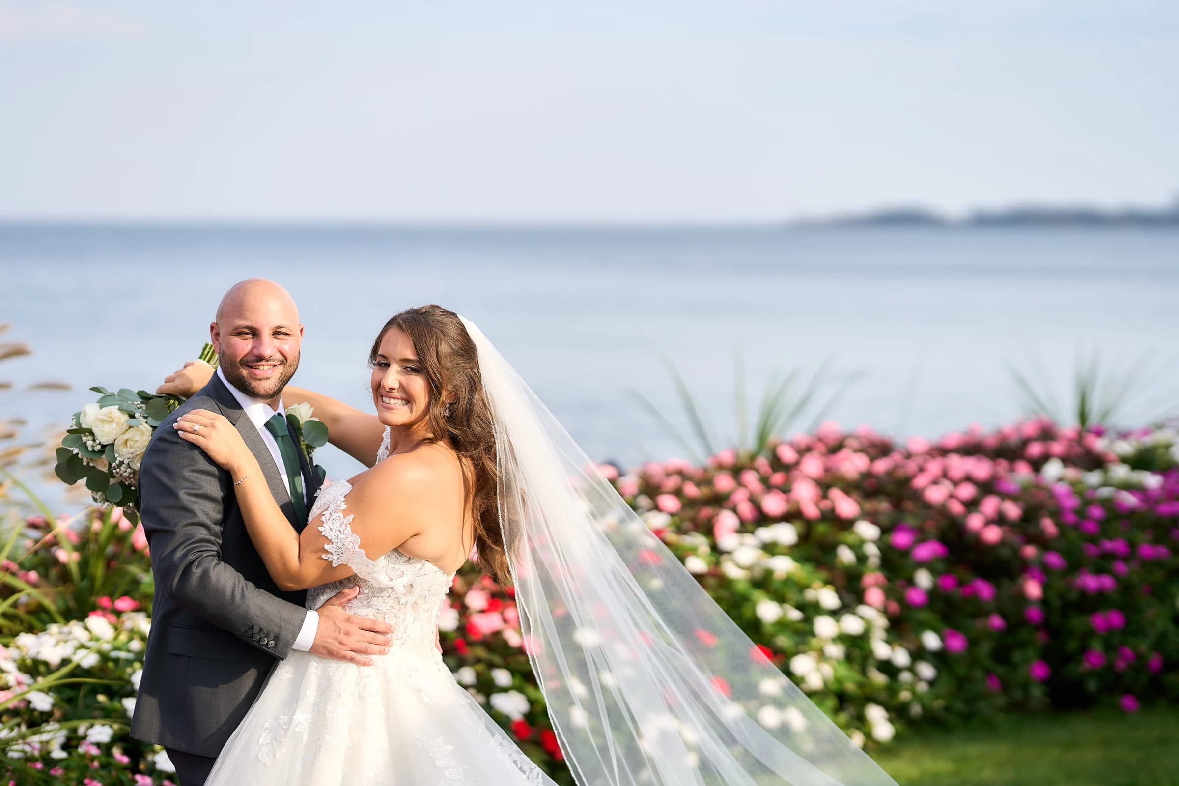 Crescent Beach Club, Bayville, Amanda and Gabriele embracing in front of the flower beds with the bay behind them