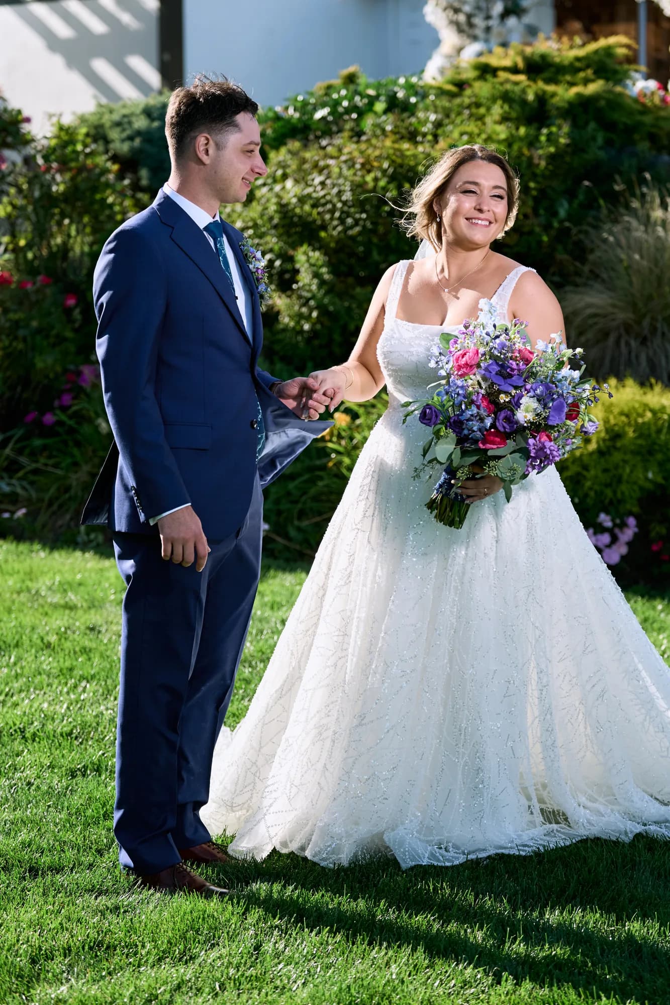 Couple backlit in warm evening light, Bride in a lace gown and groom in a navy suit share a first look in a sunlit garden