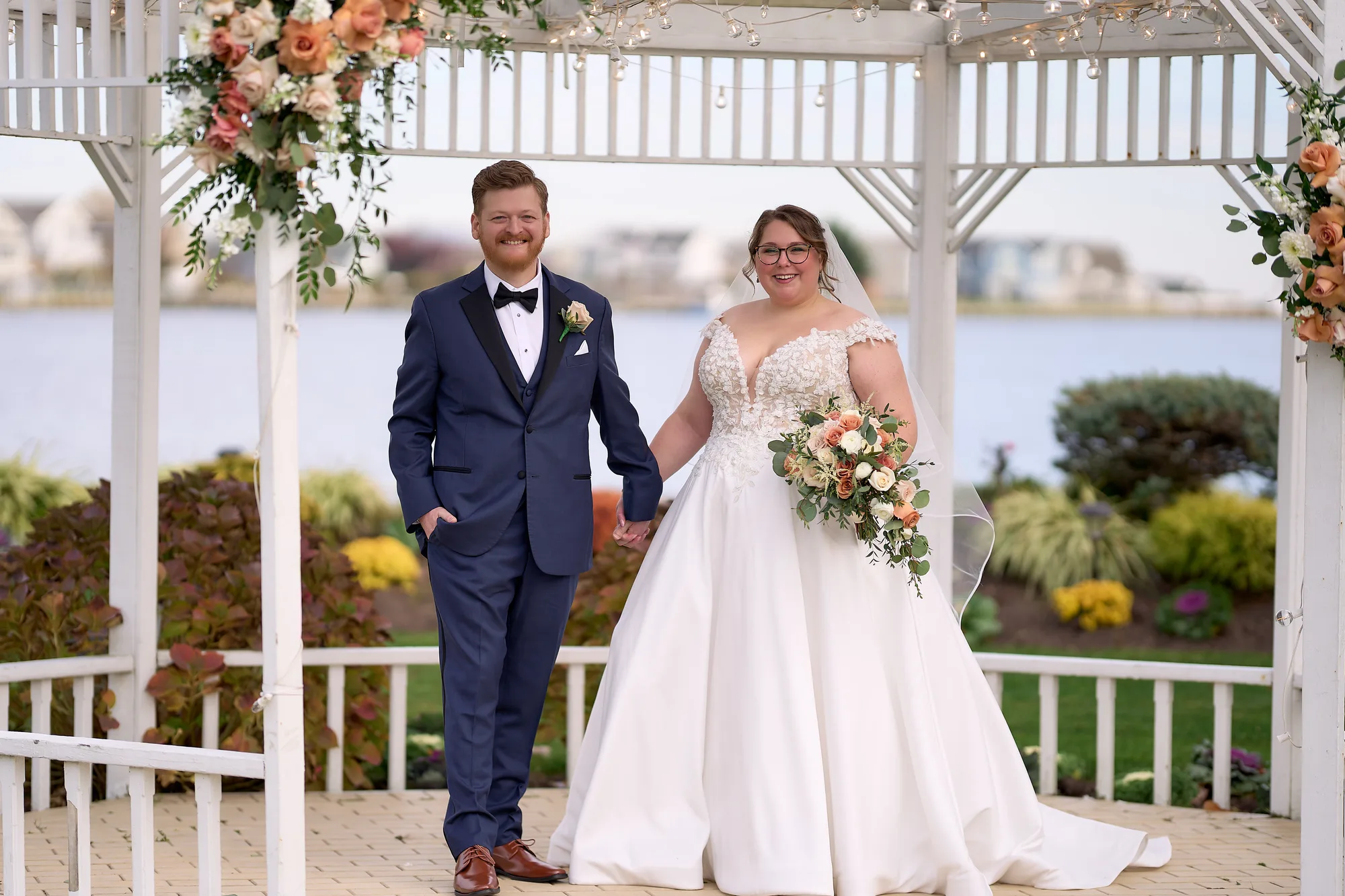 Two angles at once, the ceremony, Bride and groom holding hands under a flower-trimmed waterfront gazebo